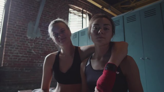 Low angle view of two female boxers with red bandaged on their hands sitting together on bench, hugging and looking at camera
