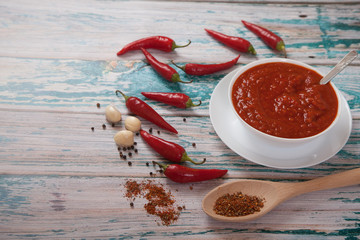 Harissa in a porcelain cup, hot red chili peppers, garlic and spices in a wooden spoon on a blue background. Flat lay. Top view.