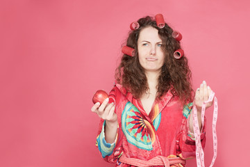 Attractive young woman with curlers in hair pours lips, holds fresh red apple, has healthy nutrition, wears red home bathrobe, stands on pink wall background, keeps to diet. People, eating concept