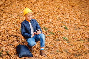 Naklejka na ściany i meble Handsome preteen boy using smartphone on a walk in autumn park. Stylish schoolboy walking after school. People, technology, fashion, lifestyle.