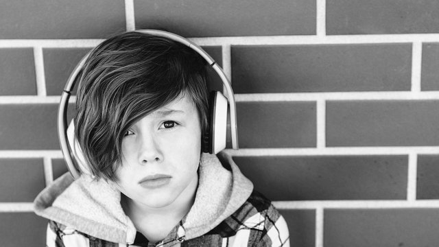 Stylish young boy in headphones listening to music over brick wall. Teenages and emotions concept. Sad handsome boy teen with headphones looking to camera.