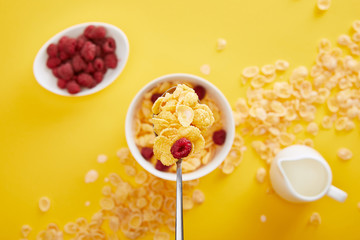 top view of spoon with cornflakes and fresh raspberry above bowl with breakfast isolated on yellow