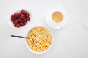 top view of bowl of cornflakes with spoon near cup of coffee and plate with raspberry on white marble surface