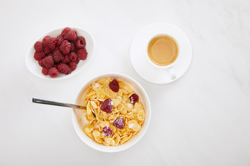 top view of bowl of cornflakes with raspberry and sugar powder near cup of coffee and plate with fresh berries on white marble surface