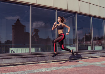 Fototapeta na wymiar Tanned woman, fitness instructor is jumping girl athlete is jogging in morning, in summer city, beautiful athlete is engaged in fitness, youth lifestyle, healthy in city. Free space for text.