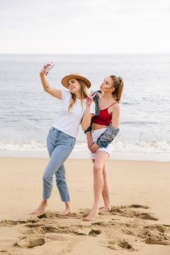 Two girls standing on sand at the beach looking at a mobile phone for a selfie