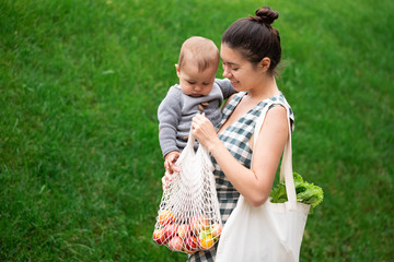 Young mother with baby son walking and shopping fruits and vegetables with reusable cotton Eco produce bag. Zero waste lifestyle concept. Concern for the next generation
