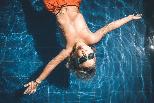 portrait of a young boy with sunglasses and colorful shorts floating in a pool