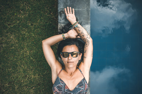 portrait from above of a tattoed black haired woman with sunglasses beside a pool
