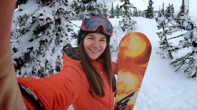 Cheerful beautiful young girl snowboarder makes selfie on an action camera or smartphone, smiling. A woman is standing among the winter snowy pines. Winter fun at the ski resort. Slow motion.