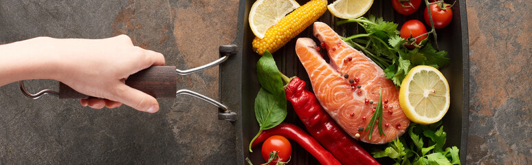 partial view of woman holding raw salmon with vegetables, lemon and herbs in grill pan, panoramic shot