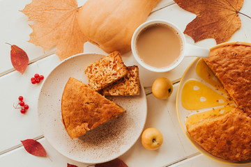 slice of pumpkin pie on a plate, a cup of coffee with milk and pumpkin on a white wooden table. dry autumn leaves and yellow apples.