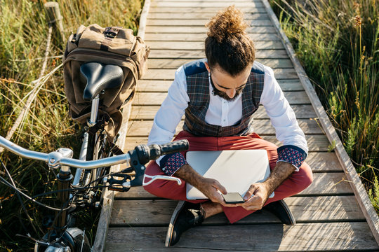 Well dressed man sitting on a wooden walkway in the countryside next to a bike with cell phone and laptop
