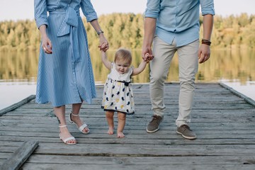 Unrecognizable stylish parents helping their baby-girl walking by the pier
