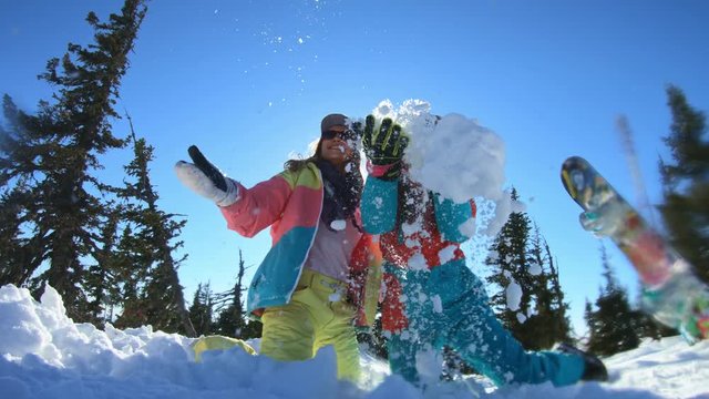 Two cheerful beautiful young girls snowboarders or skiers enjoy sitting in a snowdrift and throwing snow, smiling. Women Stands among winter snowy pines. Winter fun at the ski resort. Slow motion.