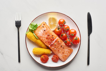 top view of raw salmon steak served with corn and tomatoes on plate near cutlery on marble table