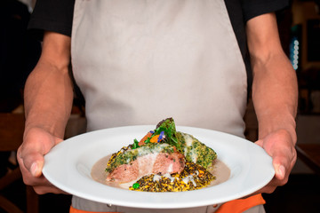 Chef holds a plate with both hands. The dish contains pepper-crusted veal with mashed sweet potato and a fresh salad over a brown sauce. Blank space on apron to place logo or brand
