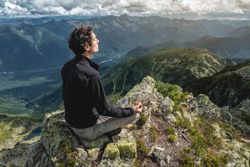 Naklejka na ściany i meble Man hiker sitting in a yoga pose at the peak of the mountain in the summer. Meditation after a long climb on a mountain