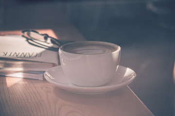 Latte coffee in white coffee cup on the wooden table in front of the large window of the coffee house.