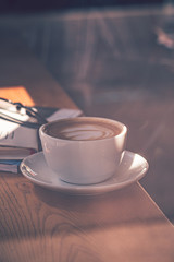 Latte coffee in white coffee cup on the wooden table in front of the large window of the coffee house.