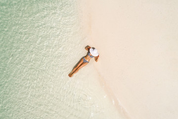 Summer holiday fashion concept - tanning girl wearing sun hat at the beach on a white sand shot from above.Top view from drone. Aerial view of slim woman sunbathing lying on a beach in Maldives.