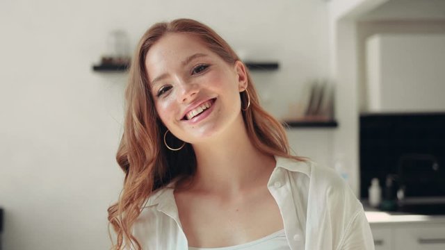 Close-up portrait of cute redhead girl with curly hair and freckles looking at camera smiling inside the room positive emotion