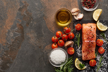 top view of raw salmon with seasoning, oil, garlic and tomatoes on stone surface