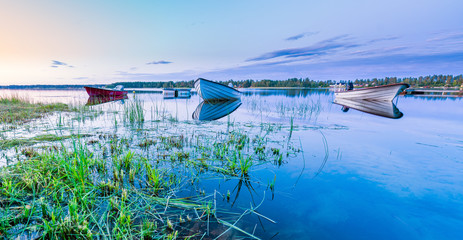 Motor and row boats anchored on the calm summer lake, sunset time, boats reflected in the water. Stocksjo Lake, close to Umea city, Northern Sweden