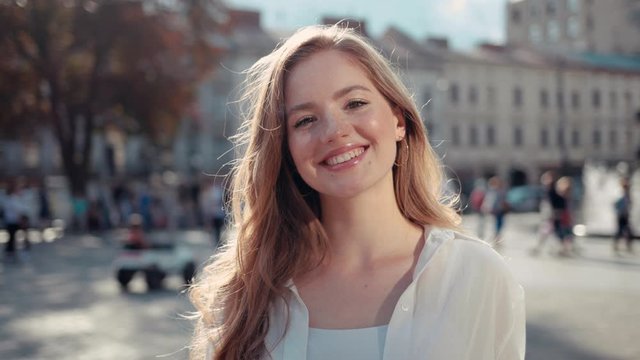 Portrait of pretty young woman with frizzy hair in city center smiling and looking at camera enjoying urban city having fun sun is shining