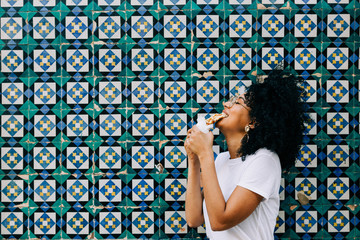 Beautiful young woman in white t-shirt standing by colorful tile wall, biting waffle