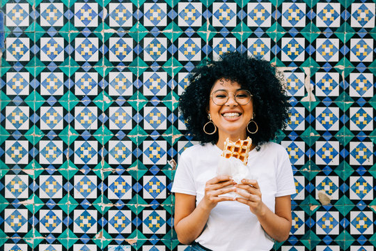 Beautiful young woman in white t-shirt standing by colorful tile wall, eating waffle and smiling at camera