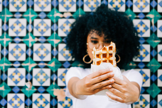 Beautiful young woman in white t-shirt standing by colorful tile wall, eating and showing waffle to camera