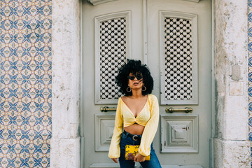 Trendy African American woman in yellow crop top and jeans standing by door and looking at camera