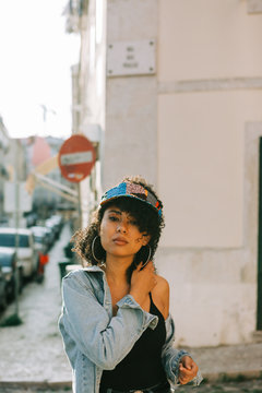 Fashionable African American woman in tank top and unbuttoned denim jacket standing on sidewalk and looking at camera
