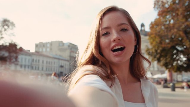 Portrait of young positive redhead woman with wavy hair in city center smiling and talking at camera making blog feel happy
