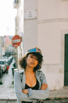 Fashionable African American woman in tank top and unbuttoned denim jacket standing on sidewalk and looking at camera