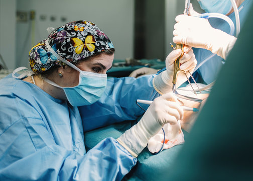 Side view of serious young doctor in protective mask and cap making surgery with instruments and crop nurse