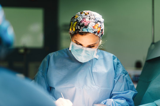 Serious young doctor in protective mask and cap making surgery with instruments and crop nurse