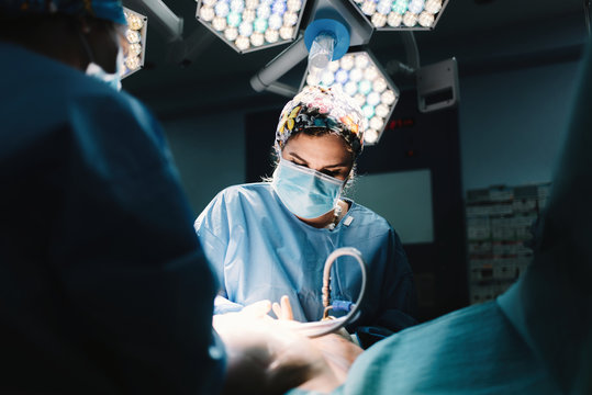 From below serious young doctor in protective mask and cap making surgery with instruments and crop nurse