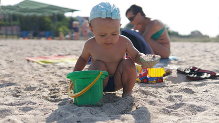 Little boy squatting on sea beach and sculpt a sand construction with pail in his hand