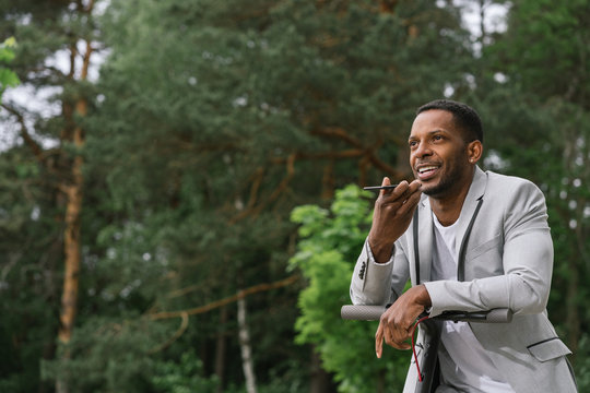 Elegant African American businessman recording voice message on mobile phone while riding on electrical scooter in park
