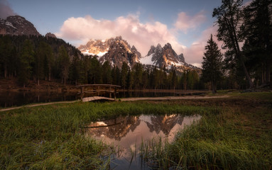 Pacifying landscape of dense forest along which path going with small bridge next to round pond spreading out on lush green lawn reflecting sky and mountains in Dolomites Itlay