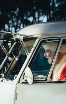 Smiling senior man in costume of Santa Claus driving old van with metal bell in hand on sunny day looking away