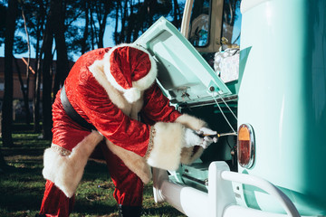 Person in red and white costume of Santa Claus inspecting engine of vintage green van on sunny day