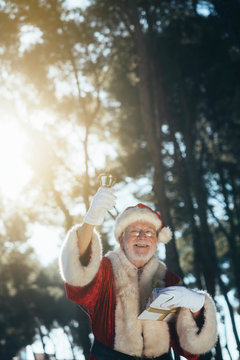 Cheerful senior man in costume of Santa Claus standing with present and bell in gloved hands looking at camera on nature background