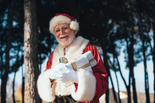 Cheerful senior man in costume of Santa Claus standing with present and bell in gloved hands looking at camera on nature background
