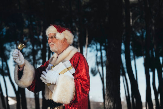 Cheerful senior man in costume of Santa Claus standing with present and bell in gloved hands looking away on nature background