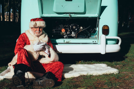 Senior man in costume of Santa Claus speaking on mobile phone while sitting by van with open engine compartment
