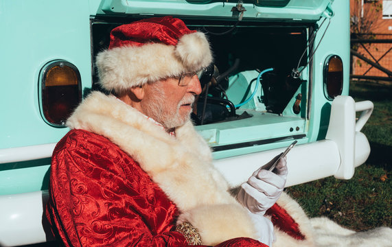 Side view of senior man in costume of Santa Claus speaking on mobile phone while sitting by van with open engine compartment