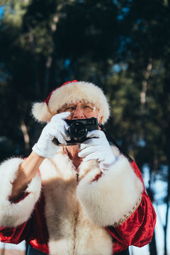 Smiling senior man in costume of Santa Claus standing and taking photos by camera on nature background looking away